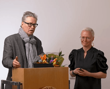 Pertti Anttonen puhujanpöntön takana, onnittelukukkakimppu, vieressä Niina Hämäläinen.