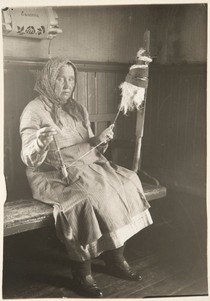 Iäkäs, huivipäinen nainen istuu vanhan talon kuluneella penkillä kehräämässä lankaa. Hän katsoo vakavana suoraan kameraan. Pään yläpuolella seinällä on kankainen lehtiteline, johon on kirjottu koristekuvioita ja sana 