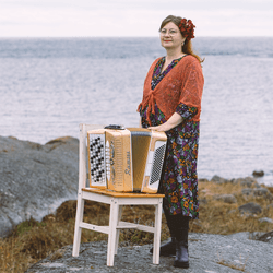 Maria Kalaniemi Utössä meren rannalla edessään tuoli, jolla on kullanvärinen 5-rivinen harmonikka.